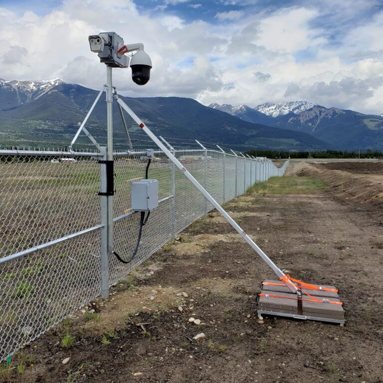 Security camera mounted on a pole with a chain-link fence in the background, overlooking a natural landscape with mountains.