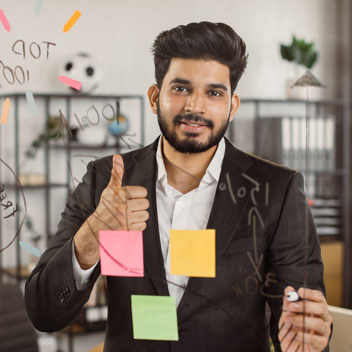 A smiling man in a business suit giving a thumbs-up in an office with sticky notes on a glass wall.