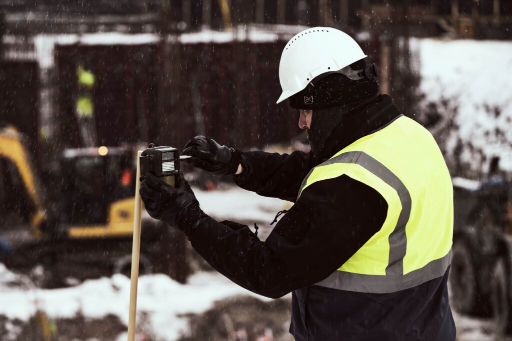 A construction worker in high-visibility clothing using surveying equipment on a snowy worksite.