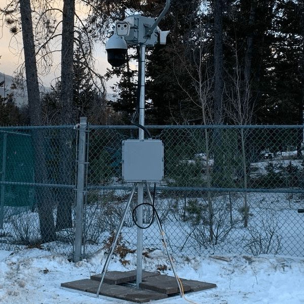 Weather monitoring station on a metal stand with electronic equipment and sensors, situated next to a chain-link fence with trees in the background during dusk.