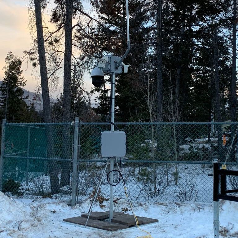 Weather monitoring station with instrumentation on a metal stand surrounded by a fence in a snowy woodland setting.