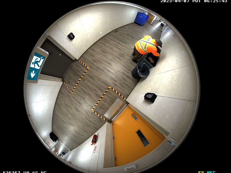 A worker in high-visibility clothing empties a trash bin in a hallway, as seen from a ceiling-mounted fisheye security camera.